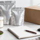 Conceptual ingredient order-planning desk with an unbranded sample jar, two sealed silver pouches, shipping carton, blank notebook, and calculator
