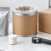 Conceptual packaging evaluation desk with an unbranded silver pouch, open lined fiber drum, shipping carton, sample jar, and handheld inspection tool
