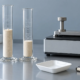 Conceptual powder-flow laboratory setup with two glass cylinders, a generic tapping-density instrument, an empty weighing dish, and a blank notebook