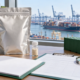 Conceptual import planning desk with sealed ingredient pouch, sample vial, document folder, clipboard, magnifying glass, and miniature cargo container overlooking a port