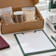 Conceptual sample evaluation desk with sealed pouches and jar in a protective box, blank clipboard, folder, digital balance, pen, and gloves